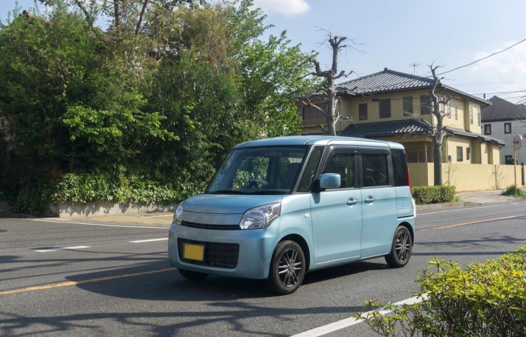 青空の中、住宅地内の公道上を走行している1台の青い軽ワゴン車