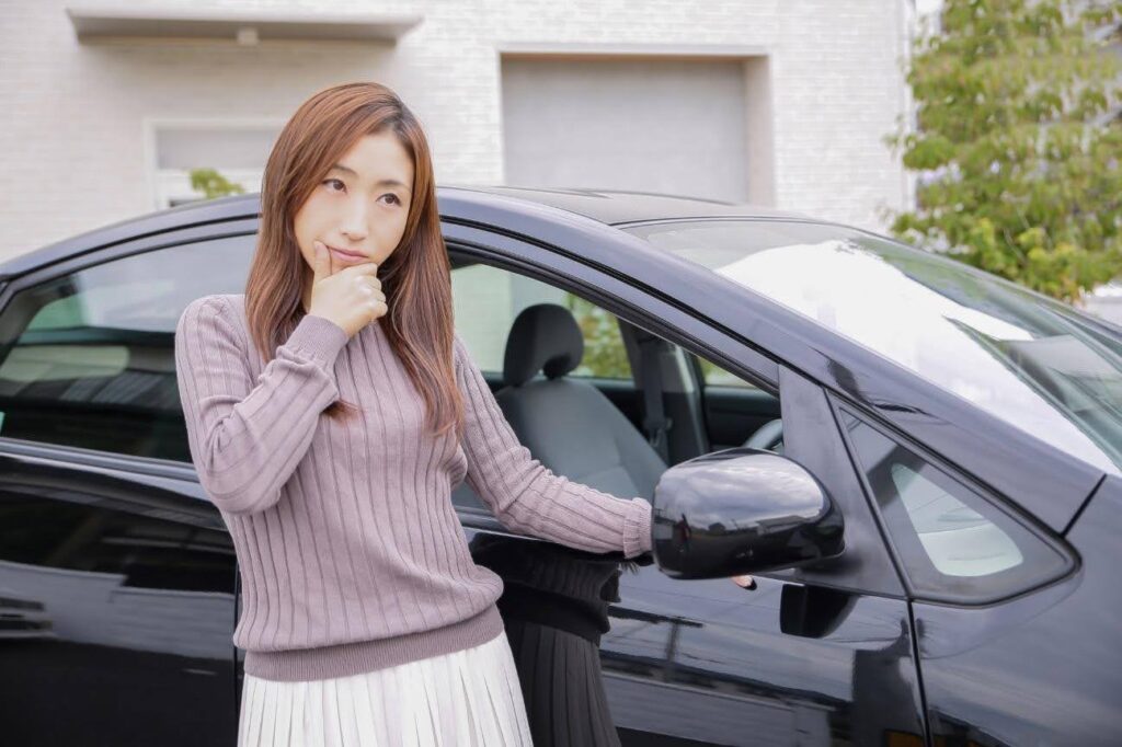 黒色の車の窓に手をかけながら顎に手を当てている茶髪の女性