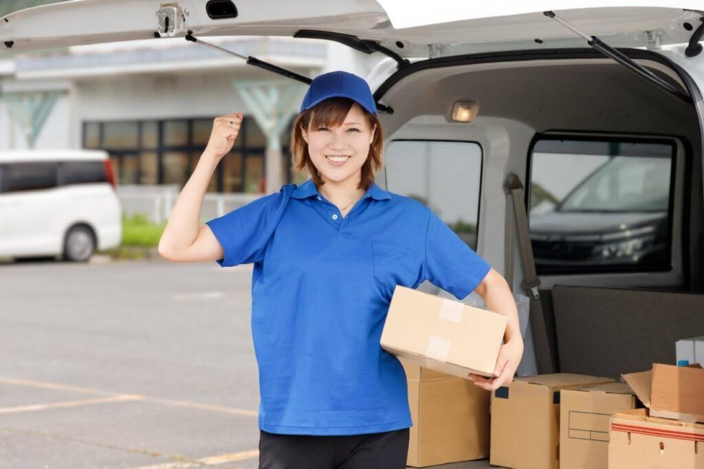 ラゲッジスペースを背に段ボールを持ちながらガッツポーズを取る青い服を着た女性